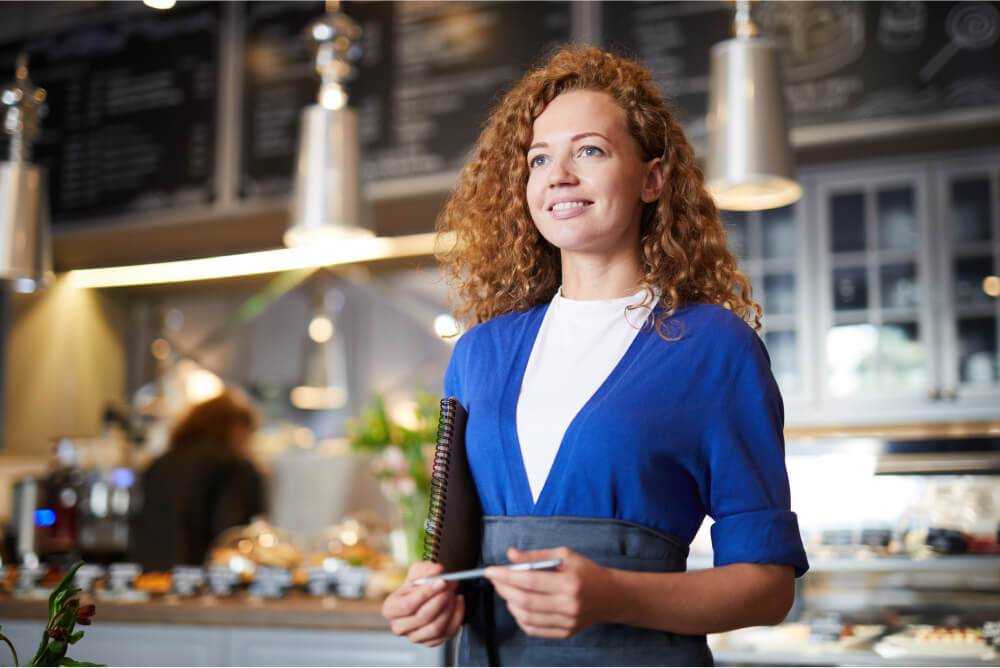 A woman with curly hair holding a notepad and pen in a cafe with a display of pastries and an Easy Solar Kit in the background.