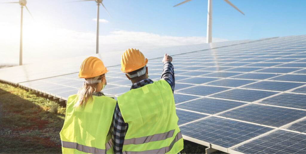 Two engineers in hard hats and reflective vests gesturing towards a wind turbine at a renewable energy site, discussing the installation of an Easy Solar Kit.