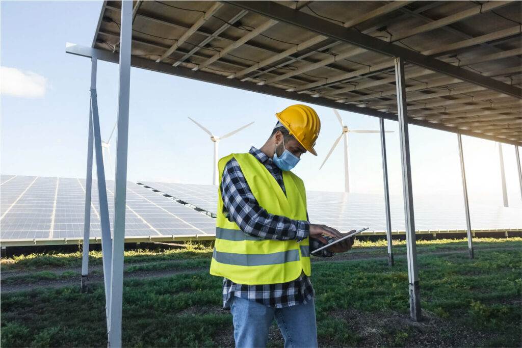 Engineer with tablet inspecting a solar farm equipped with an Easy Solar Kit and wind turbines in the background.