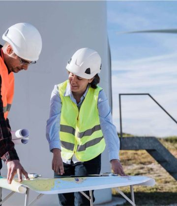 Two engineers in safety vests and hardhats discussing plans for a Micro Inverter installation in front of a wind turbine.